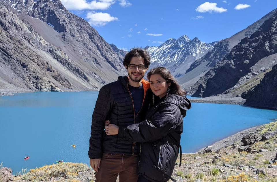 Casal posando abraçado em Portillo, com a Laguna del Inca azul ao fundo e montanhas nevadas da Cordilheira dos Andes sob céu azul.