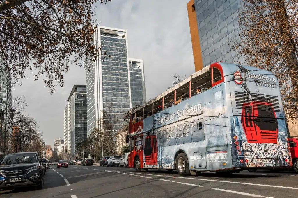 Ônibus turístico hop-on-hop-off em avenida de Santiago, cercado por prédios altos e árvores secas em dia claro.