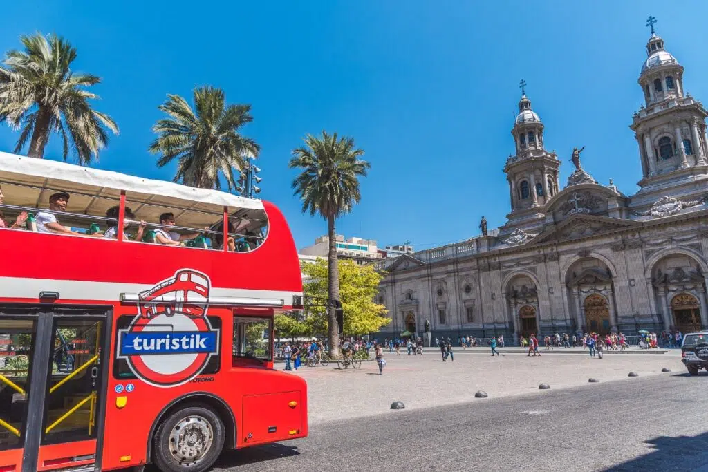 Ônibus vermelho de dois andares do city em Santiago hop-on-hop-off em frente à Catedral Metropolitana, com palmeiras e céu azul ao fundo.