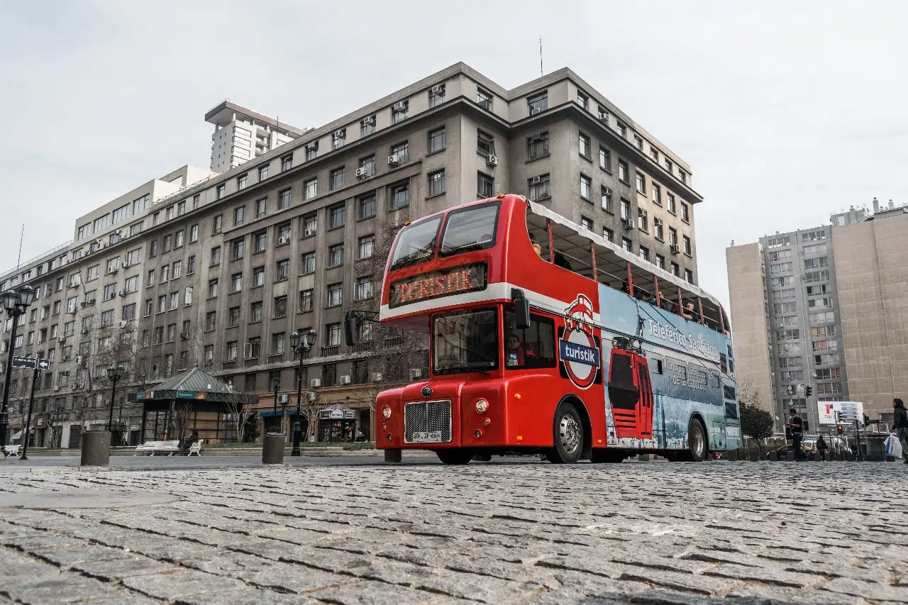 Ônibus vermelho de dois andares do tour hop-on-hop-off em Santiago, estacionado em praça urbana cercada por prédios, em dia nublado.
